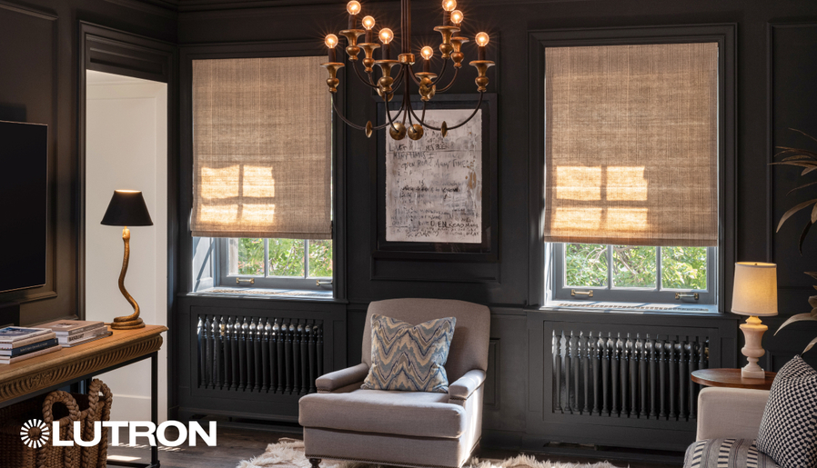 A cozy, elegant living room with dark walls features motorized Lutron shades partially drawn over tall windows, a classic chandelier, and a plush armchair centered below framed artwork.