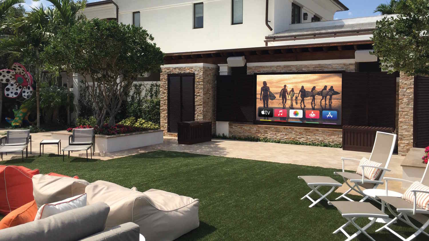 Large outdoor TV mounted on a stone wall in a landscaped backyard with lounge seating, creating a luxury al fresco entertainment space.