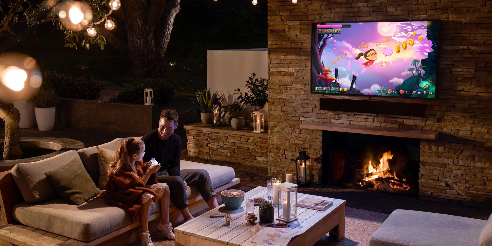 Father and daughter enjoying a game on an outdoor TV mounted above a stone fireplace in a cozy evening patio setting.