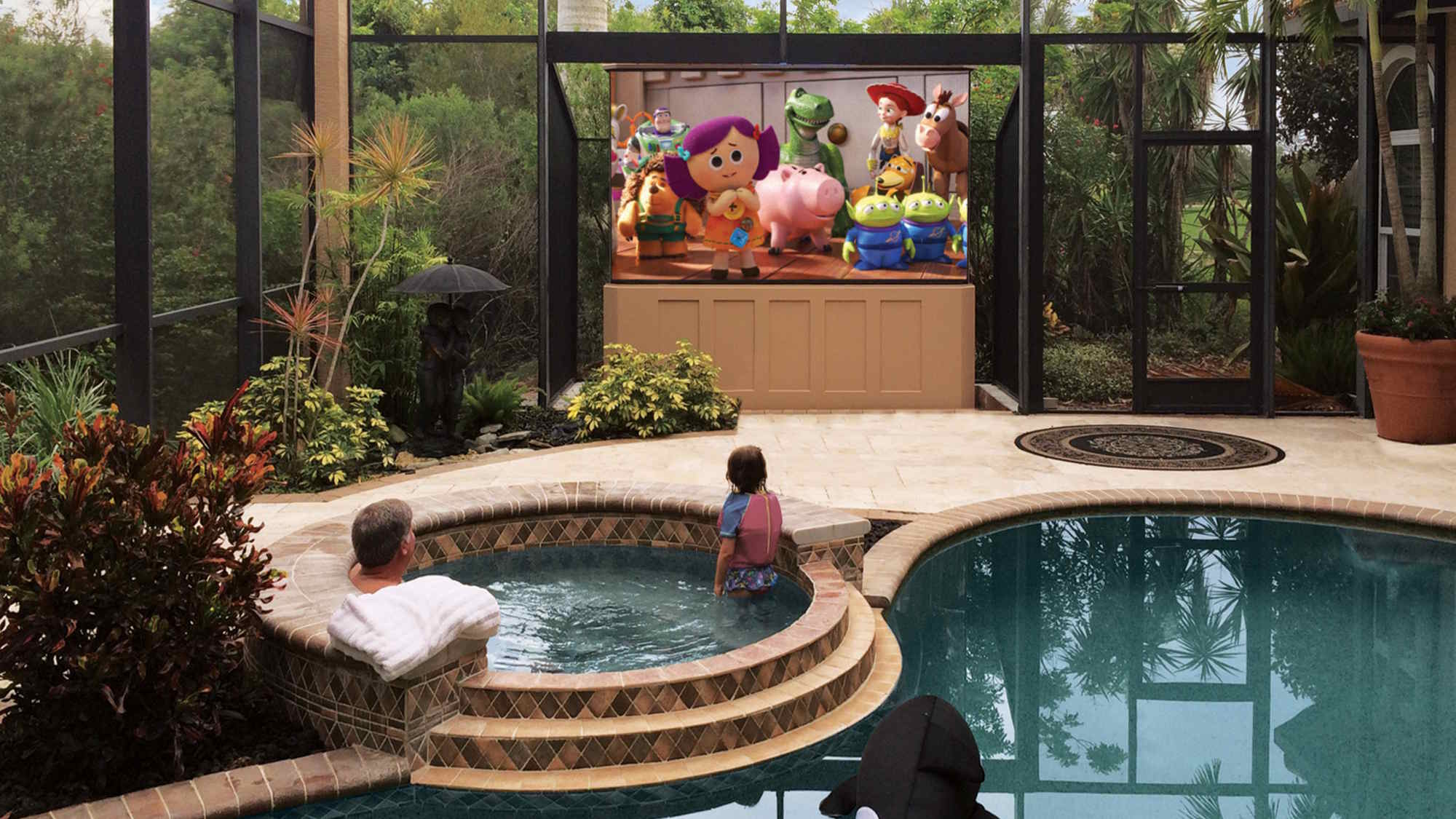 Family enjoying a movie on a large outdoor TV from a poolside spa in a screened lanai, showcasing weatherproof outdoor entertainment.