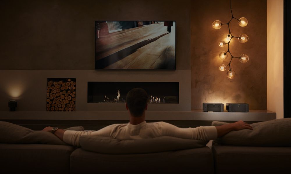 <span class="uk-h4">Person sits on couch looking at a large TV display on the wall. The room is dimly lit for enhanced viewing.</span>