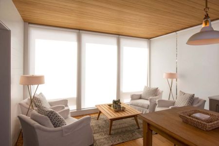 A bright and cozy living room with four white armchairs arranged around a wooden coffee table, set against tall windows with sheer roller shades and a wood-paneled ceiling.