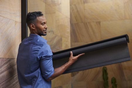 A man in a blue shirt holding a rolled-up black window shade, standing in front of a stone-tiled wall with a cactus in the background.