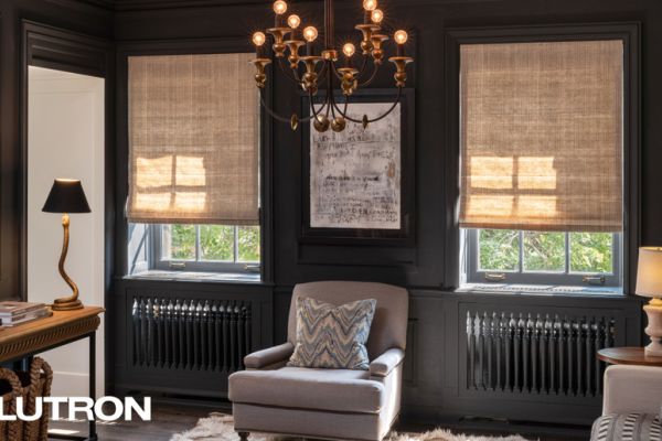 A cozy, elegant living room with dark walls features motorized Lutron shades partially drawn over tall windows, a classic chandelier, and a plush armchair centered below framed artwork.
