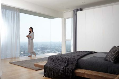 A minimalist bedroom with a low-profile wooden bed, dark gray bedding, floor-to-ceiling windows offering a panoramic city view, and a woman in a robe standing by the sheer curtains holding a cup.