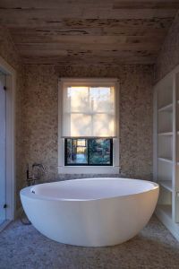A serene bathroom with a large freestanding oval bathtub positioned beneath a window covered by a partially lowered roller shade, surrounded by textured stone walls and wooden ceiling panels.